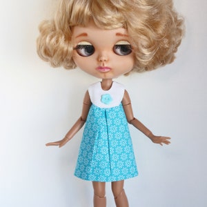 May include: A doll with blonde curly hair wearing a blue and white floral dress with a white collar and a light blue flower button.