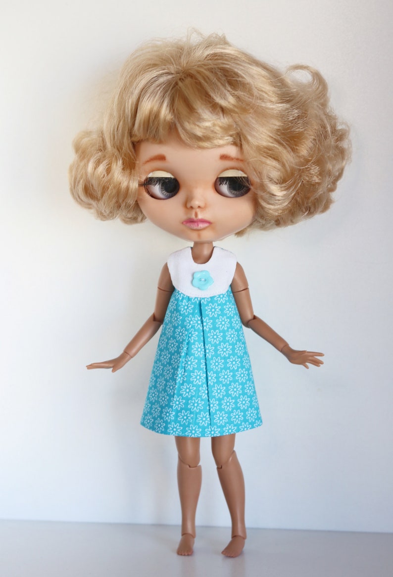May include: A doll with blonde curly hair wearing a blue and white floral dress with a white collar and a light blue flower button.