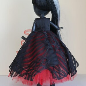May include: A fashion doll with black hair and a silver streak, wearing a black dress with red accents. The dress features a layered skirt with a striped pattern and a red tulle underskirt. The doll is posed from the back.