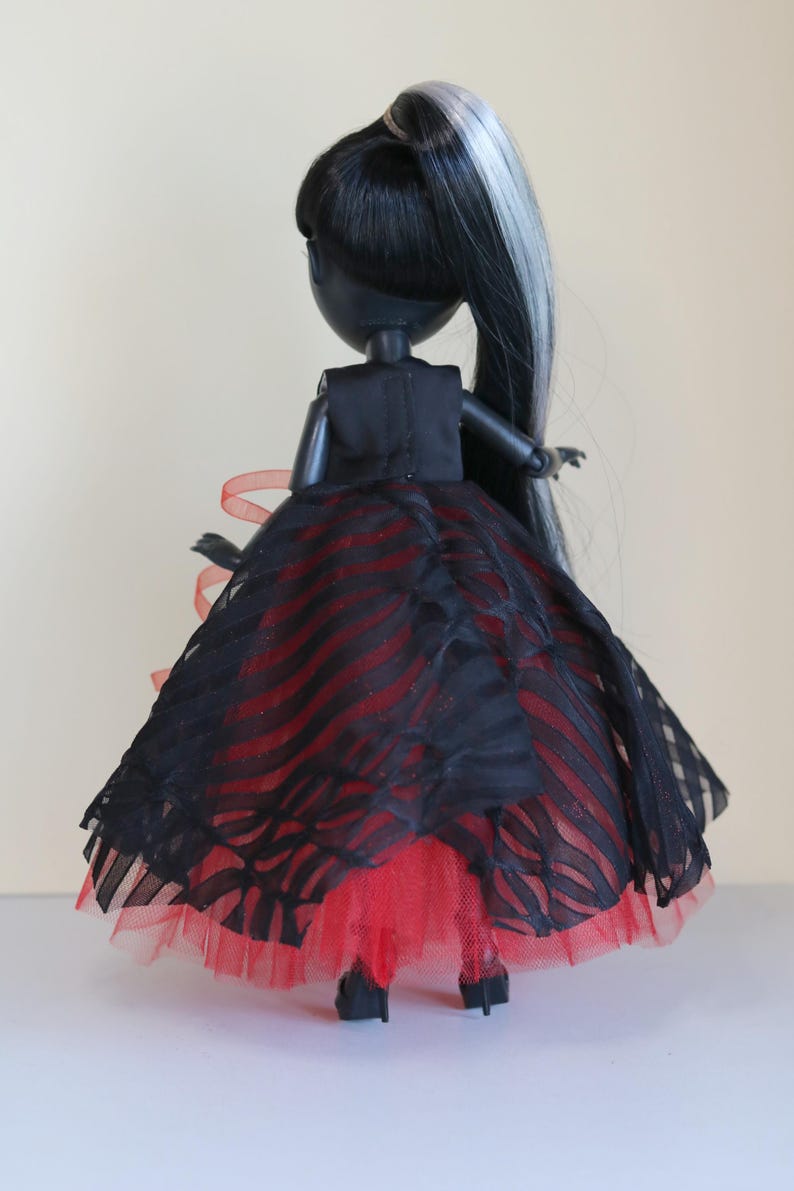 May include: A fashion doll with black hair and a silver streak, wearing a black dress with red accents. The dress features a layered skirt with a striped pattern and a red tulle underskirt. The doll is posed from the back.