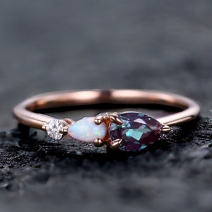 May include: A rose gold ring with a pear-shaped alexandrite gemstone, a pear-shaped opal, and a small round diamond. The ring is set on a dark background.
