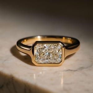 Op de afbeelding: Een gouden ring met een rechthoekige, heldere edelsteen. De edelsteen is gezet in een bezel setting en de band is glad en gepolijst. De ring wordt gepresenteerd op een marmeren oppervlak.