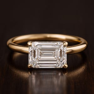 May include: A gold ring with a large, rectangular-cut diamond. The clear, multifaceted diamond is set in a simple, polished gold band. The ring is presented on a dark, reflective surface.