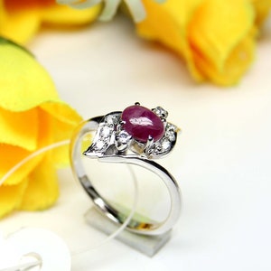 May include: A silver ring with a large, oval-cut ruby gemstone. The ruby is a deep red colour and is surrounded by smaller, clear gemstones. The ring has a leaf-like design on one side, with a polished finish.