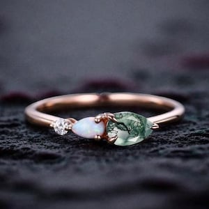 May include: A rose gold ring with three gemstones. A small, round, clear gemstone is on the left, followed by a white oval stone, and a pear-shaped green stone on the right. The ring is set against a dark background.