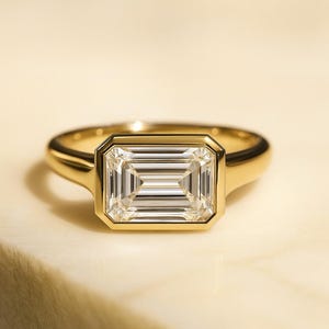 Op de afbeelding: Een gouden ring met een rechthoekige smaragdgeslepen diamant. De diamant is gezet in een bezel-zetting en de band is glad, gepolijst goud. De ring wordt getoond op een lichtgekleurd oppervlak.