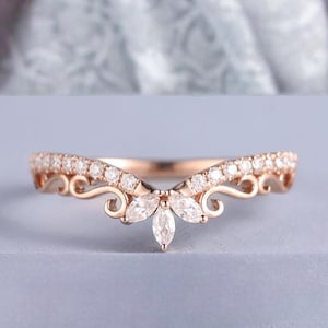 Puede incluir: Una delicada banda de boda de oro rosa con un diseño en forma de V y un grupo de diamantes de forma marquesa en el centro.