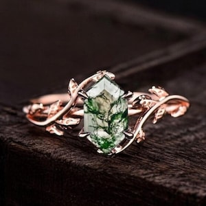 Puede incluir: Anillo de oro rosa con una gema de ágata musgosa verde facetada. El anillo tiene un diseño inspirado en la naturaleza con detalles de hojas en la banda. La gema es de color verde claro con inclusiones verdes más oscuras.