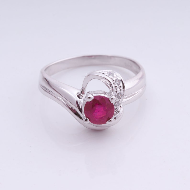 May include: A silver ring featuring a round, deep red gemstone at the center, accented by smaller, clear stones. The ring has a unique, flowing design with a curved band and a decorative swirl.