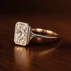 May include: A radiant-cut diamond ring with a gold band. The rectangular diamond is set in a bezel setting, showcasing its facets. The ring is displayed on a dark wooden surface, highlighting the gold and diamond's brilliance.