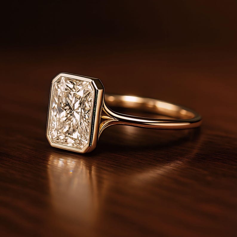 May include: A radiant-cut diamond ring with a gold band. The rectangular diamond is set in a bezel setting, showcasing its facets. The ring is displayed on a dark wooden surface, highlighting the gold and diamond's brilliance.