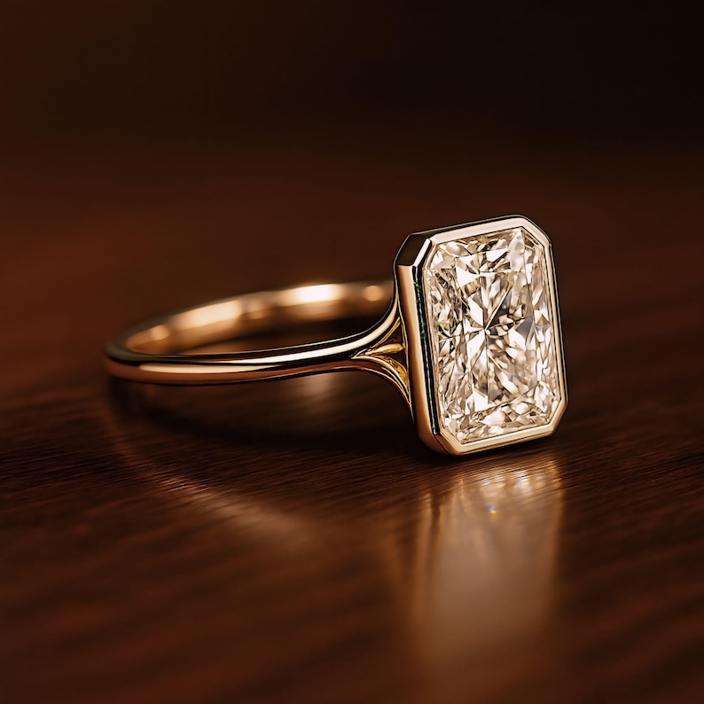 May include: A gold ring with a large, rectangular diamond. The diamond is set in a bezel setting, and the band is a simple, polished gold. The ring is displayed on a dark wooden surface, reflecting the light.