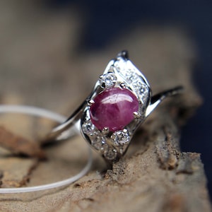 May include: A silver ring with a central oval ruby, a deep red hue. The ring is embellished with smaller, clear gemstones. The design is elegant, with the ruby as the main focus, complemented by the surrounding smaller stones.