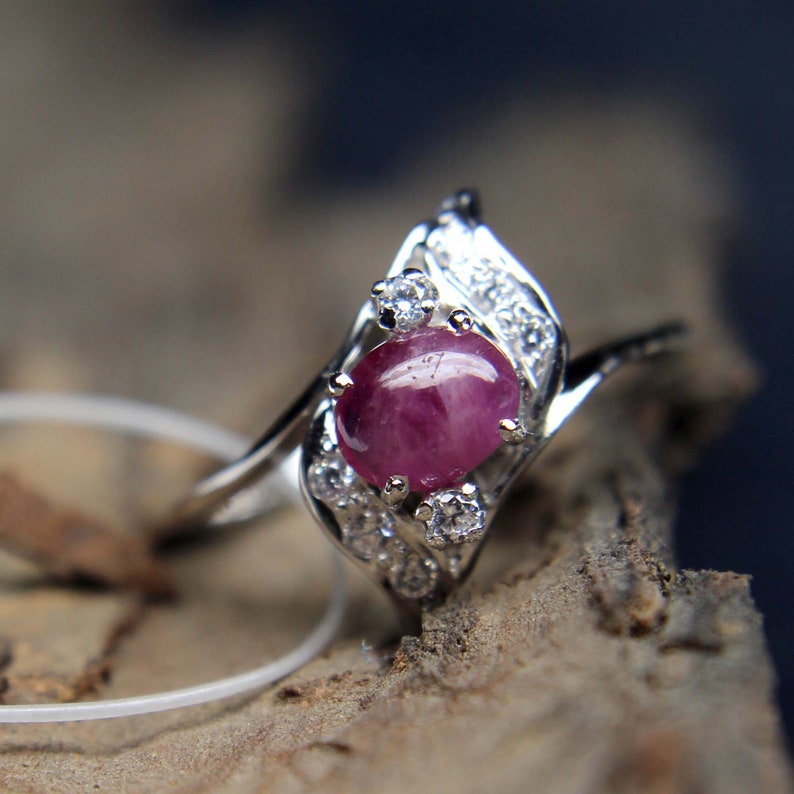 May include: A silver ring with a central oval ruby, a deep red hue. The ring is embellished with smaller, clear gemstones. The design is elegant, with the ruby as the main focus, complemented by the surrounding smaller stones.