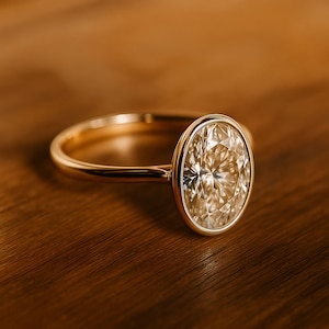 May include: A gold ring featuring an oval-cut diamond. The diamond is set within a bezel. The ring is displayed on a wooden surface. The ring is a piece of jewellery.