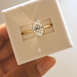 May include: A gold ring featuring an oval-cut diamond in a bezel setting. The ring is presented in a white box. The diamond is clear and the gold band is slender and polished. A timeless piece of jewellery.