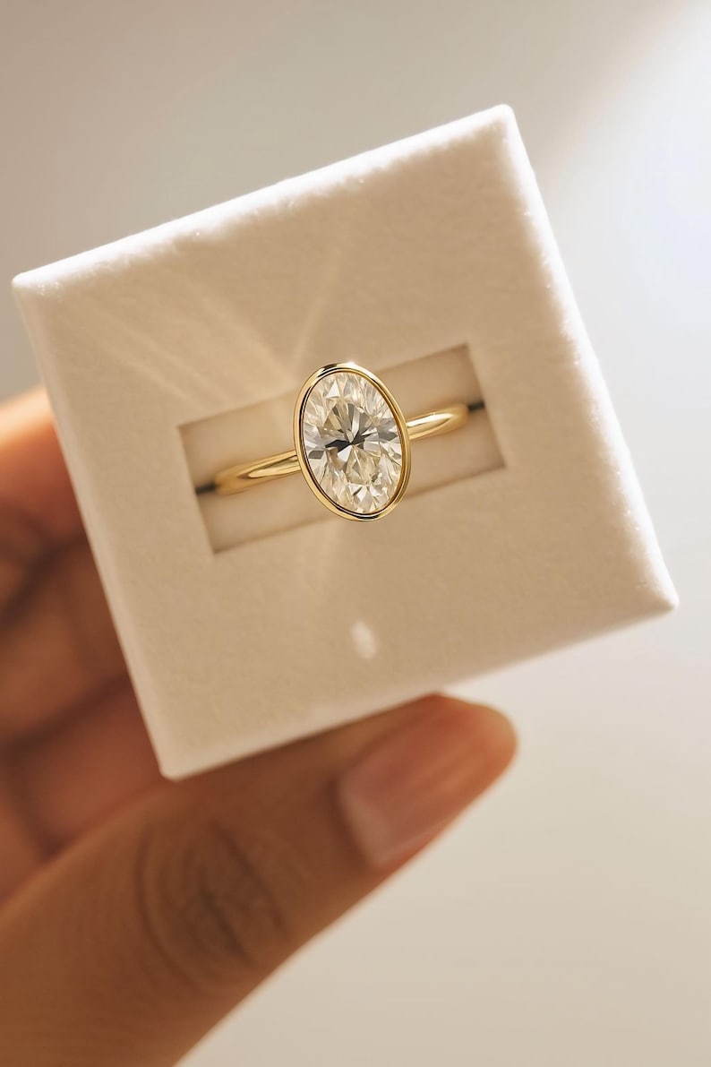 May include: A gold ring featuring an oval-cut diamond in a bezel setting. The ring is presented in a white box. The diamond is clear and the gold band is slender and polished. A timeless piece of jewellery.