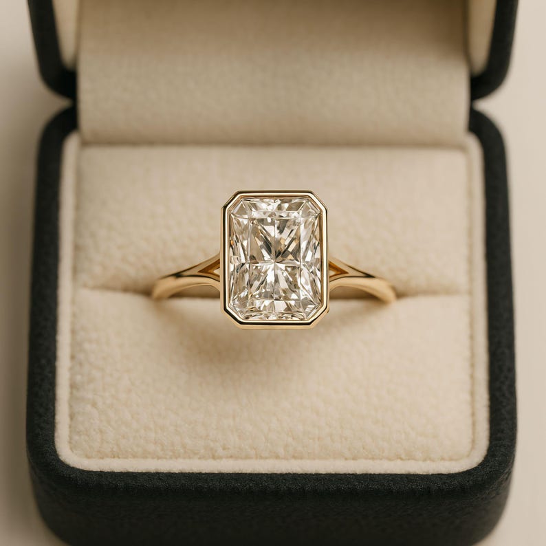 May include: A gold ring with a large, rectangular, clear gemstone set in a bezel setting. The ring is displayed in a black ring box with a cream-colored interior. The band of the ring is a delicate gold color.