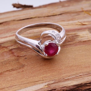 May include: A silver ring with a large, round, deep red ruby as the primary stone. The ring has a unique design with a curved band and small accent diamonds. The ring is set against a wooden background.