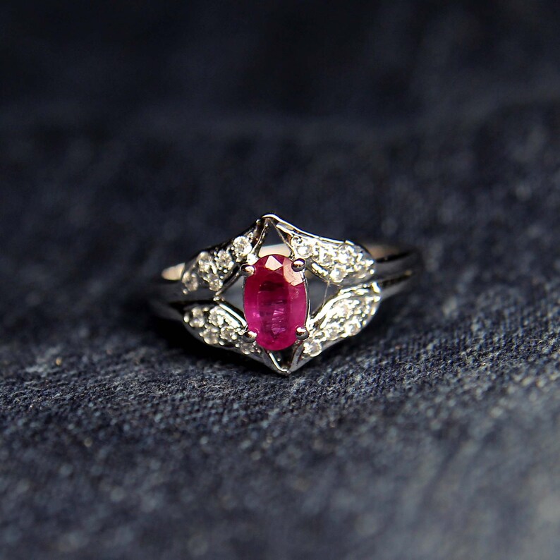 Puede incluir: Un anillo de plata con una piedra preciosa ovalada de rub&iacute; rojo rodeada de peque&ntilde;as piedras blancas.