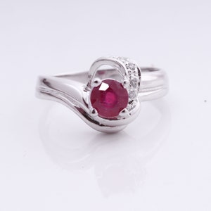 May include: A silver ring with a large, round, deep red gemstone as the focal point. The ring features a swirling design with small, clear accent stones. The band has a smooth, polished finish, creating a classic and elegant look.