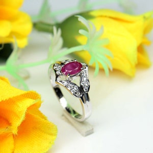 Puede incluir: Un anillo de plata con una gran piedra de rub&iacute; ovalada rodeada de diamantes m&aacute;s peque&ntilde;os. El anillo est&aacute; colocado sobre un fondo blanco con flores amarillas.