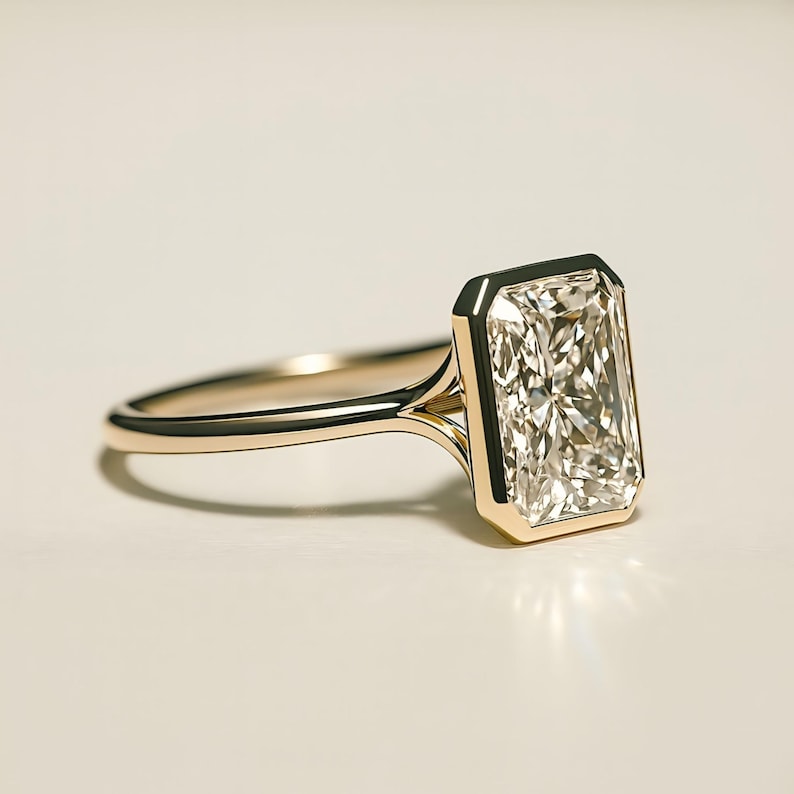 May include: A gold ring with a large, rectangular, clear gemstone. The gemstone is set in a bezel setting, and the band is a simple, polished gold. The ring is displayed on a white surface.