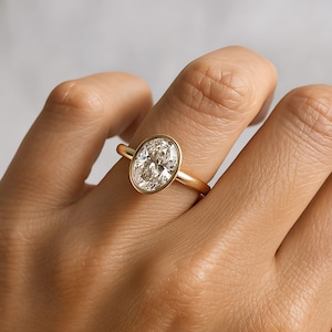 May include: A gold ring with an oval-cut diamond. The diamond is set in a bezel setting, and the band is plain, polished gold. The ring is displayed on a finger, showcasing the design and sparkle of the gemstone.