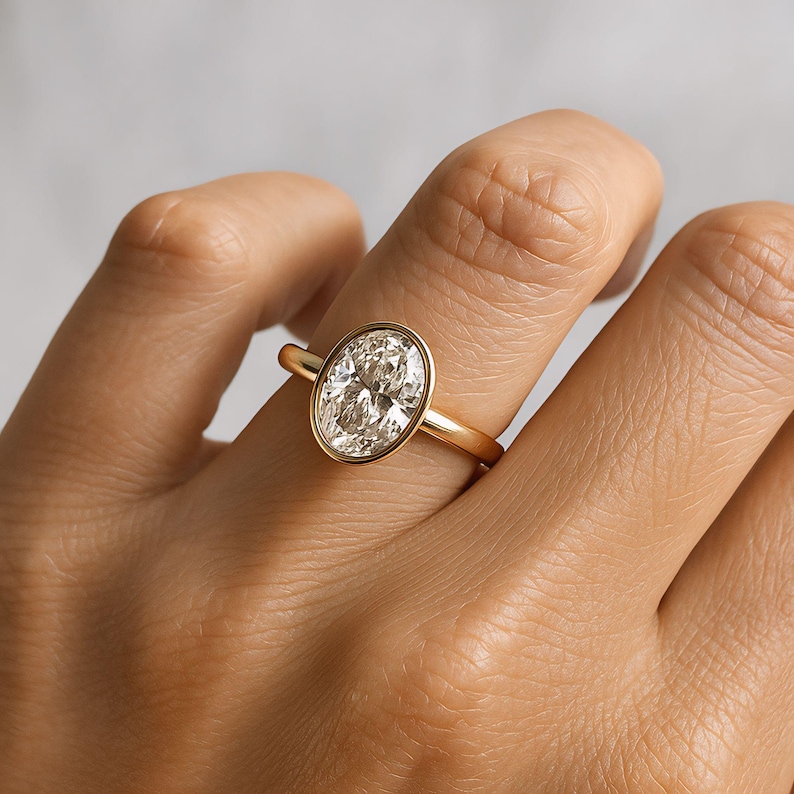 May include: A gold ring with an oval-cut diamond. The diamond is set in a bezel setting, and the band is plain, polished gold. The ring is displayed on a finger, showcasing the design and sparkle of the gemstone.