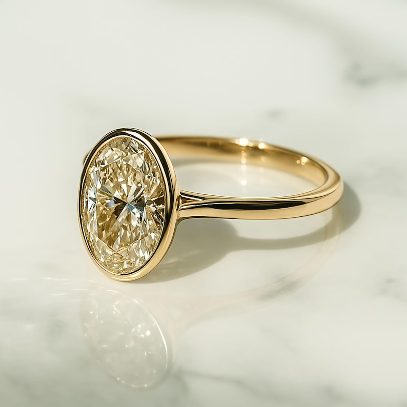 May include: A gold ring with an oval-cut diamond. The diamond is set in a bezel setting, and the band is polished gold. The ring is shown on a white marble surface, reflecting light. The ring is a piece of jewellery.