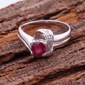 May include: A silver ring with a round, deep red gemstone as the focal point. The ring features a decorative setting with small, clear accent stones. The band has a smooth, polished finish.