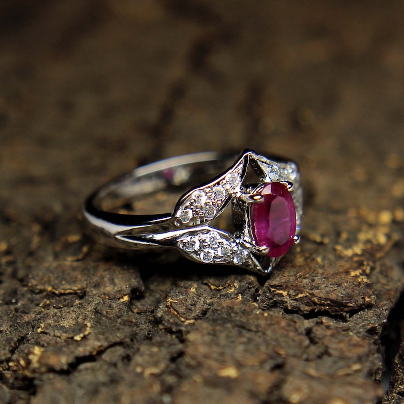 Puede incluir: Un anillo de plata con una gran piedra de rub&iacute; ovalada rodeada de piedras peque&ntilde;as transparentes. El anillo tiene un delicado dise&ntilde;o floral.