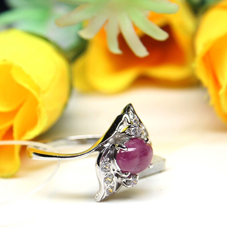 May include: A silver ring with a large, oval, deep pink gemstone, likely a ruby, set in an ornate setting. The ring is embellished with small, clear gemstones. The design incorporates a floral or leaf-like pattern.