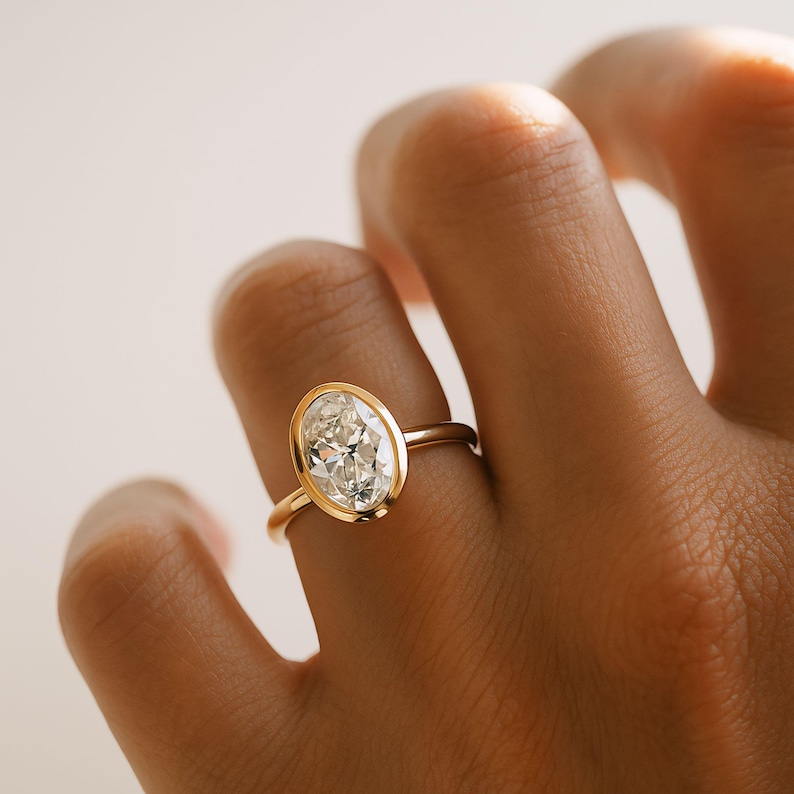 May include: A gold ring featuring a large, oval-shaped diamond. The diamond is set within a bezel, and the band is plain, polished gold. The ring is on a finger.