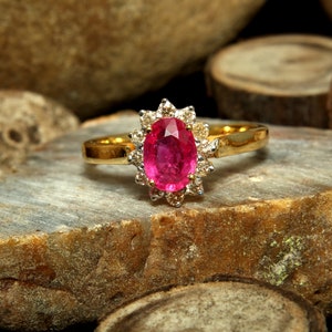 May include: A gold ring featuring a large, oval-cut ruby gemstone surrounded by a halo of smaller diamonds. The ruby is a vibrant pinkish-red color, and the diamonds sparkle against the gold band. The ring is set on a natural stone surface.