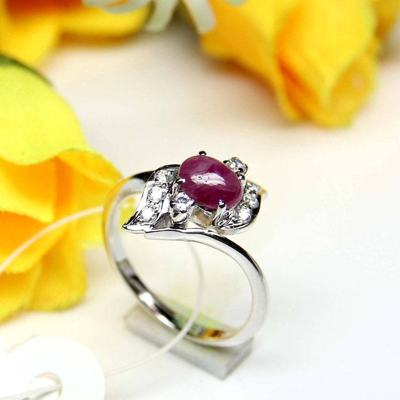 May include: A silver ring with a large, oval-cut ruby gemstone. The ruby is a deep red colour and is surrounded by smaller, clear gemstones. The ring has a leaf-like design on one side, with a polished finish.