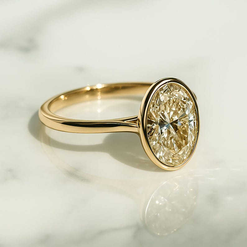 May include: A gold ring featuring an oval-cut diamond. The diamond has a light yellow hue and is set within a bezel. The polished band is smooth. The ring is presented on a white surface.