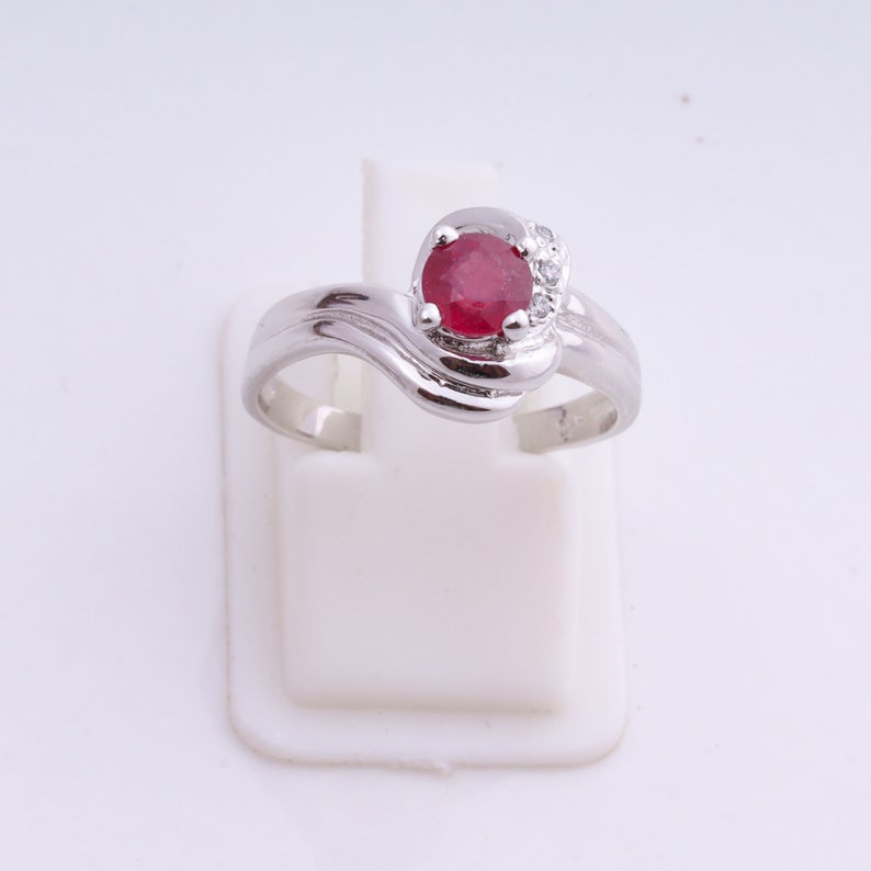 May include: A silver ring with a large, round, red gemstone as the focal point. The gemstone is surrounded by smaller, clear stones. The ring has a swirling, wave-like design on the band.