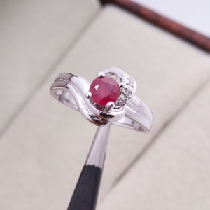 May include: A silver ring with a large, round, red gemstone as the focal point. The ring features a swirling design on the band and is accented with small, clear gemstones. The ring is held by a pair of tweezers.