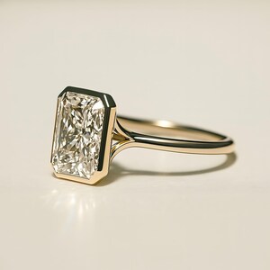 May include: A radiant-cut diamond ring with a gold band. The rectangular diamond is set in a bezel setting, showcasing its brilliance. The gold band has a delicate, streamlined design, creating a classic and elegant look. The ring is a symbol of luxury and commitment.