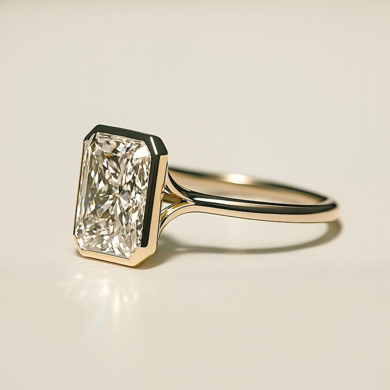 May include: A radiant-cut diamond ring with a gold band. The rectangular diamond is set in a bezel setting, showcasing its brilliance. The gold band has a delicate, streamlined design, creating a classic and elegant look. The ring is a symbol of luxury and commitment.