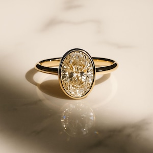 May include: A gold ring with an oval-cut diamond. The diamond is set in a bezel setting, and the band is plain gold. The ring is shown on a white surface, with a shadow behind it. The ring is a piece of jewellery.