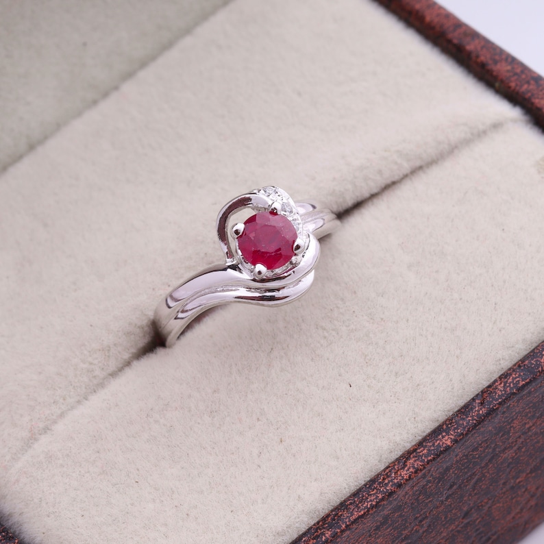 May include: A silver ring with a large, round, deep red gemstone at the center. The ring has a swirling design around the gemstone, with small, clear accent stones. The ring is set in a light-colored jewelry box.