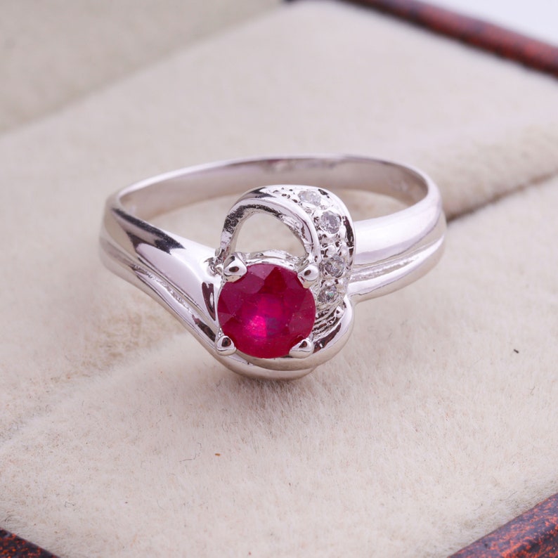 May include: A silver ring featuring a vibrant, round, red gemstone as the focal point. The ring has a unique, flowing design with a small cluster of clear stones accenting the gemstone. The band curves gracefully, creating an elegant and eye-catching piece of jewelry.