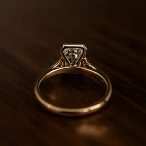 May include: A gold engagement ring with a large, clear, emerald-cut diamond. The ring's band is a simple, polished gold, and the diamond is set in a bezel setting. The ring is displayed on a dark wooden surface.