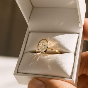 May include: A gold ring featuring an oval-cut diamond in a bezel setting, presented in a white ring box. The ring has a slender, polished band. The diamond has a pale yellow tint and is the central element of the image.