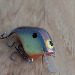 Puede incluir: Un señuelo de pesca colorido con un labio de plástico transparente. El señuelo presenta un diseño azul, naranja y amarillo con un ojo negro y dos anzuelos triples. El señuelo tiene el texto "Kerman 26" escrito.