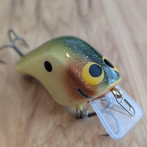 Puede incluir: Un señuelo de pesca con un cuerpo amarillo, verde y naranja, con detalles de ojos negros y un labio de plástico transparente. El señuelo tiene dos anzuelos triples y está diseñado para atraer peces. El señuelo está sobre una superficie de madera.