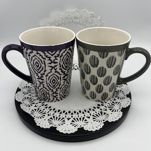 May include: Two ceramic coffee mugs with different geometric patterns. The mug on the left has a purple rim and a white and purple geometric pattern. The mug on the right has a gray rim and a white and gray polka dot pattern.