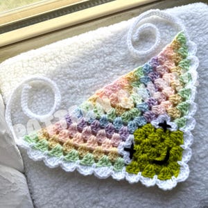 May include: A crocheted rainbow bandana with a white border and a green smiley face in the center.